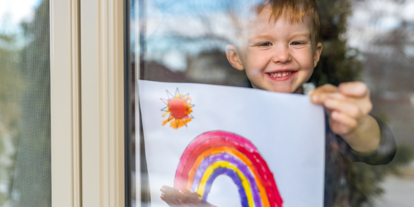 young boy holding a picture of a rainbow up to a window