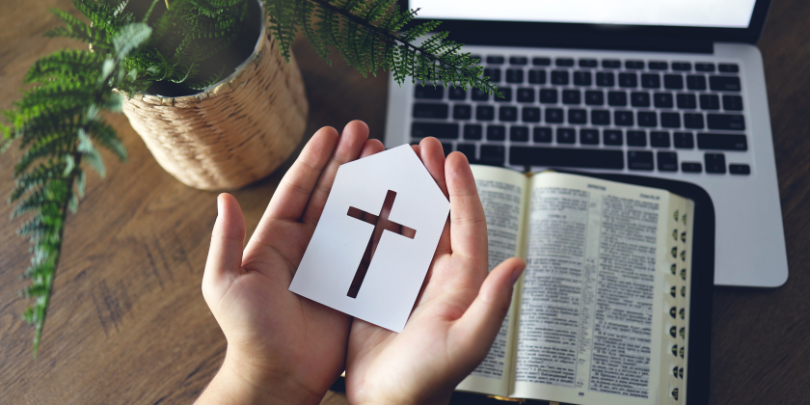 photo of someone holding the outline of a church with a cross over a bible and laptop