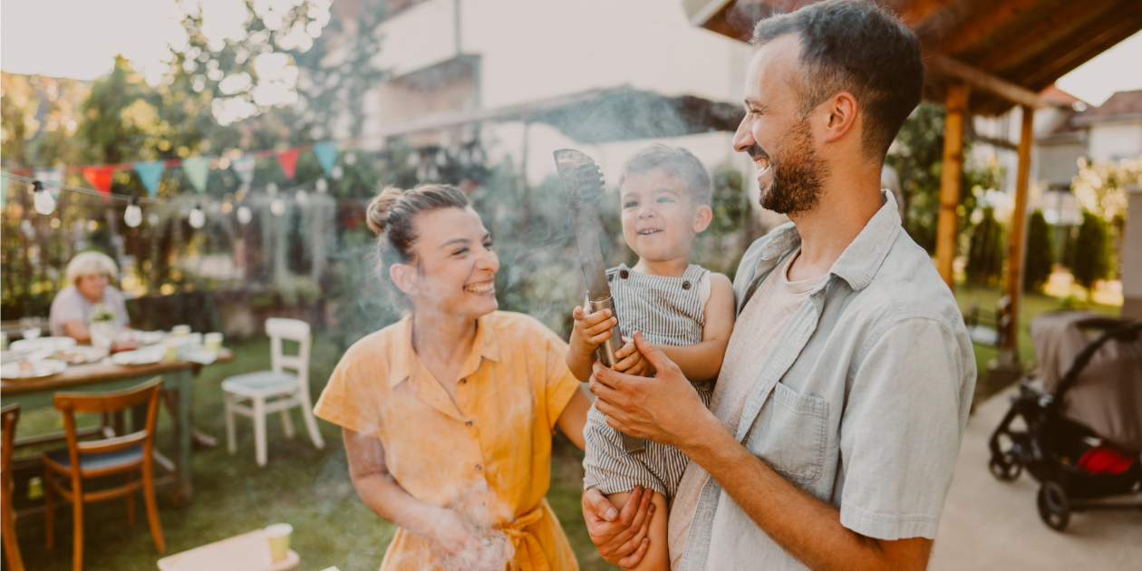 photo of parents holding a child in front of smoking grill turning corn on the cob